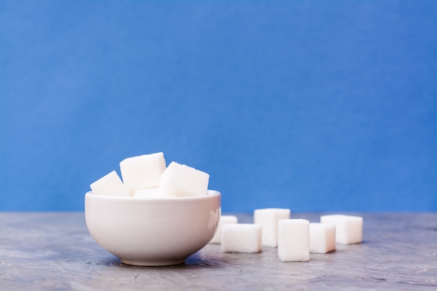 Cubes de sucre dans un bol blanc et à côté de la table sur un fond bleu