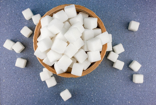 Cubes de sucre dispersés dans un bol en bois sur fond bleu Sucre dans un bol en bois sculpté sur un gros plan de table bleu