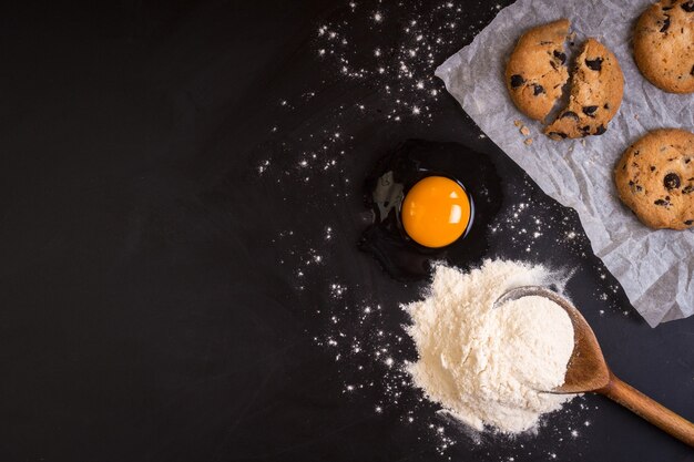 Cuillère en bois rustique avec une farine, un œuf cru, un papier sulfurisé et des biscuits sur un tableau noir