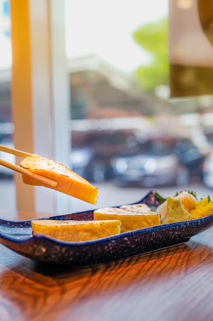 Cuisine japonaise, Focus sur un morceau de Tamago ou une omelette grillée roulée qui a été choisie par des baguettes et d'autres morceaux dans une petite assiette bleue sur une table en bois