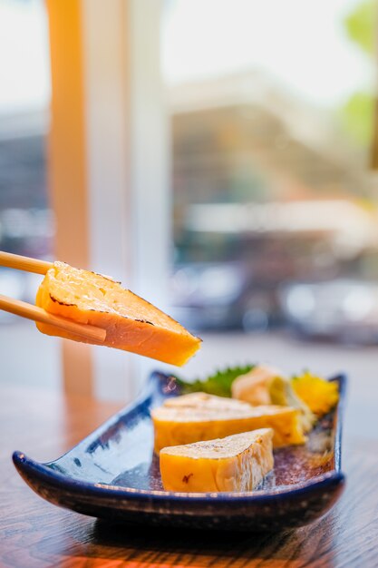 Cuisine japonaise, se concentrer sur un morceau de Tamago ou une omelette roulée grillée qui a été choisie par des baguettes et d'autres morceaux dans une petite assiette bleue sur une table en bois