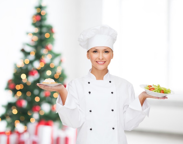 cuisine, personnes, vacances et concept alimentaire - chef féminin souriant, cuisinier ou boulanger tenant un petit gâteau et une salade sur des assiettes et montrant un geste correct sur le salon avec fond d'arbre de noël