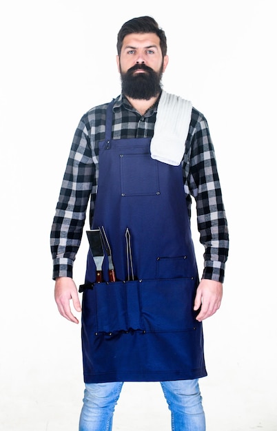 Cuisine en plein air Homme barbu portant un tablier avec des outils de préhension du gril dans les poches Hipster avec des ustensiles en métal pour le gril du barbecue Cuisinier au gril Préparer des aliments sur le gril à l'aide d'un ensemble de barbecue