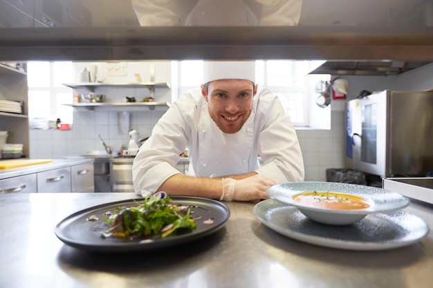 cuisine, profession et concept de personnes - chef cuisinier heureux avec une assiette de soupe et de salade à la table de cuisine d'un restaurant