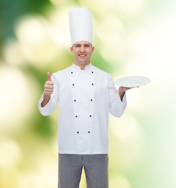 cuisine, profession, publicité et concept de personnes - heureux chef cuisinier tenant quelque chose sur une assiette vide et montrant les pouces vers le haut sur fond vert