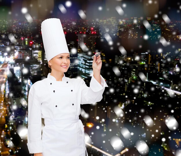 Photo cuisine, vacances, publicité et concept de personnes - chef féminin souriant, cuisinier ou boulanger avec un marqueur écrivant quelque chose sur un écran virtuel sur fond de ville de nuit enneigée