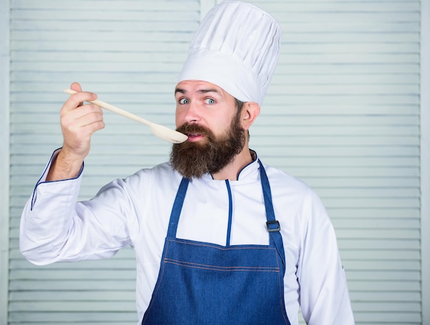 Cuisiner comme occupation professionnelle Le chef barbu hipster tient une cuillère en bois Ustensiles de cuisine et concept de cuisine Essayons de goûter Ajoutez quelques épices Homme avec barbe en chapeau de cuisinier et tablier tenir des ustensiles de cuisine