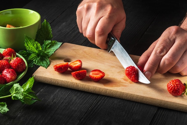 Cuisiner des desserts diététiques. Les mains du chef en gros plan ont coupé des fraises fraîches sur une planche à découper de la cuisine du restaurant pour faire des boissons gazeuses à la menthe