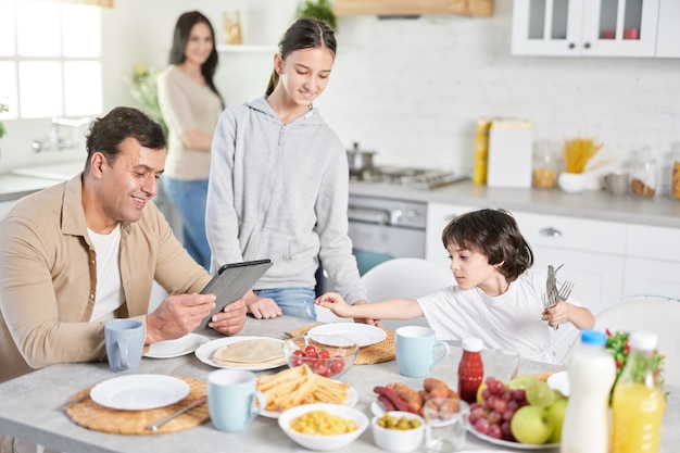 Cuisinez Pour Servir L'amour. Mari Latin D'âge Moyen Utilisant Une Tablette, Regardant Les Informations, Attendant Le Dîner Pendant Que Ses Enfants Et Sa Femme Servent Une Table Dans La Cuisine à La Maison