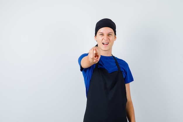 Cuisinier adolescent masculin en t-shirt, tablier pointant vers l'avant et semblant joyeux, vue de face.