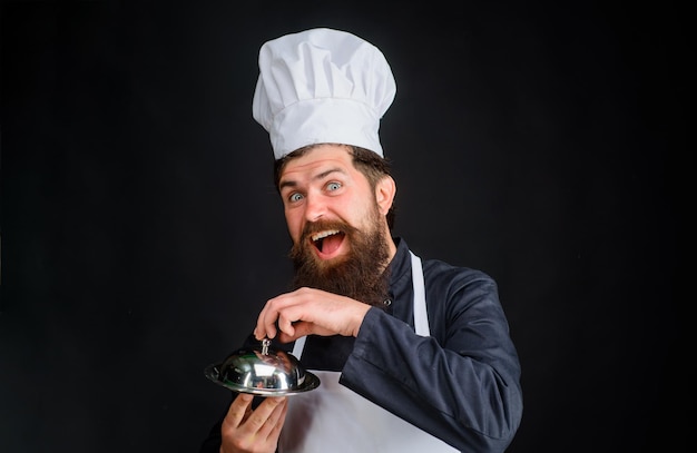 Le cuisinier de cuisine tient le plat métallique servant et le chef de présentation le cuisinier tient le chef barbu cloche tient