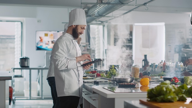 Cuisinier masculin analysant une recette culinaire sur une tablette numérique dans la cuisine, utilisant Internet sur un gadget pour préparer la nourriture. Chef professionnel dans un repas gastronomique de cuisine uniforme à partir d'un site Web de gastronomie en ligne.