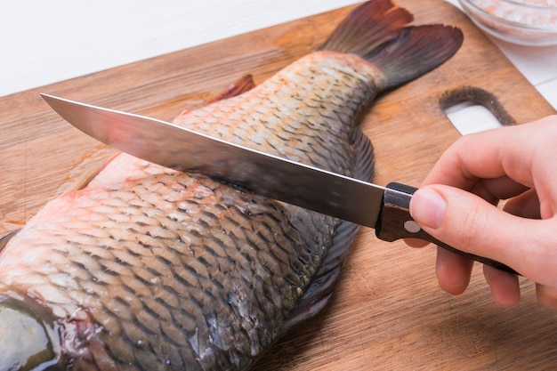 Le cuisinier nettoie les écailles de la carpe fraîche sur une planche à découper en gros plan