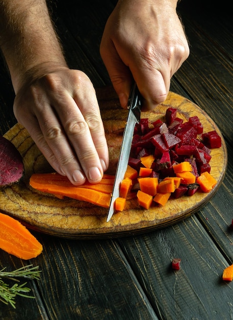 Le cuisinier prépare une vinaigrette à partir de légumes bouillis en tranchant des carottes et des betteraves avec un couteau sur une planche à couper en bois.