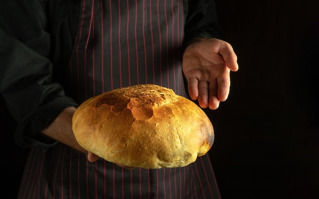 Le cuisinier présente du pain de blé fraîchement cuit dans sa main Place pour la publicité sur un fond sombre Le concept de cuisson du pain rond