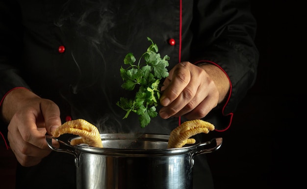 Un cuisinier en uniforme noir ajoute du persil à une casserole de pattes de poulet bouillantes Préparer une délicieuse soupe ou un plat d'accompagnement gras dans la cuisine Concept de cuisine avec poulet ou coq sur fond noir