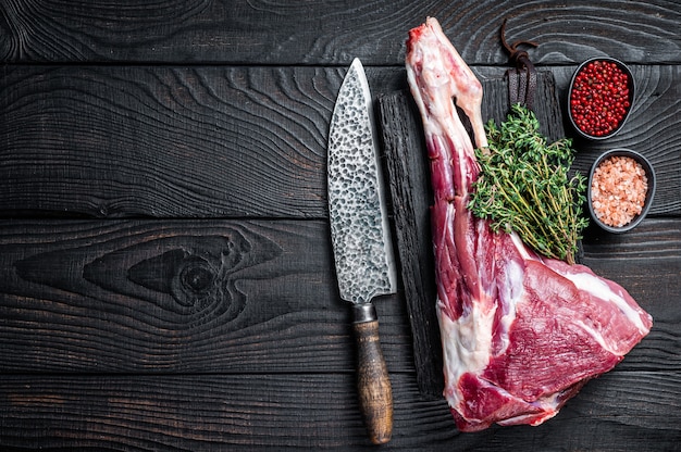 Cuisse de mouton d'agneau cru non cuit aux herbes sur une planche à découper de boucher. Fond en bois noir. Vue de dessus. Espace de copie.