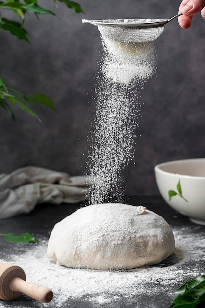 Cuisson du pain maison au levain. Tamiser la farine sur la pâte à pain crue sur la table, fond sombre, mise au point sélective.