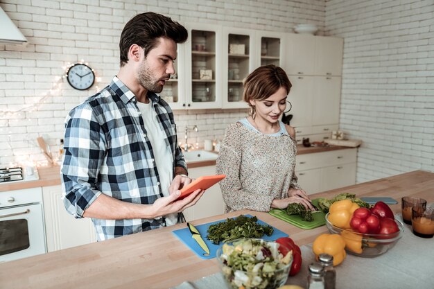 Cuisson du petit déjeuner. Jeune jolie femme avec de grandes boucles d'oreilles à la satisfaction tout en coupant les verts dans la cuisine