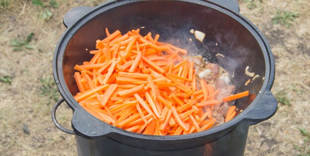 Cuisson du pilaf dans un wok en fonte à l'air libre. Beaucoup de carottes tranchées et de viande frites dans un wok sur une cuisinière avec un feu ouvert, dîner en famille à l'extérieur, gros plan.