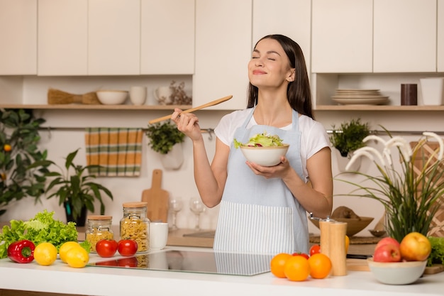 Cuisson jeune femme dans la cuisine avec une cuillère en bois. Essaye la nourriture. Concept de blogueur alimentaire. Souriant.