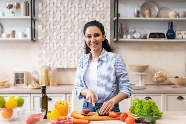 Cuisson à la maison femme latine heureuse préparant une salade de légumes dans une cuisine moderne coupant le concombre et
