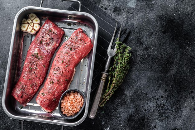 Cuisson de la viande de filet de filet d'agneau aux herbes et à l'huile d'olive, viande de mouton marinée dans un plateau de cuisine. Fond noir. Vue de dessus. Espace de copie.