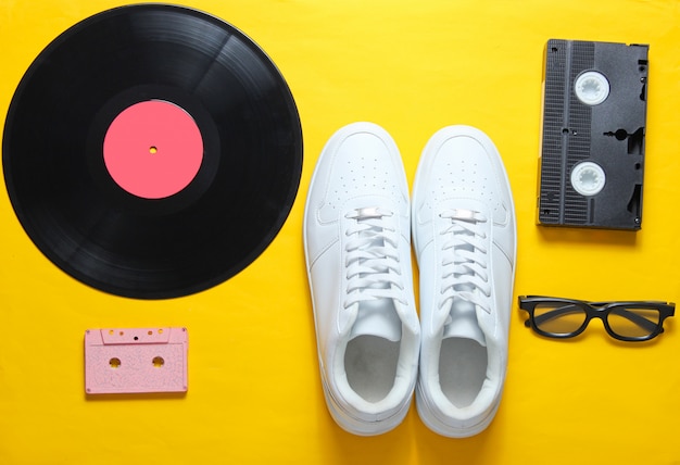 Culture pop. Baskets blanches hipster, plaque vinyle, cassette audio et vidéo, lunettes 3d sur fond jaune.