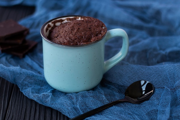 un cupcake au chocolat dans une tasse est cuit au micro-ondes Mise au point sélective En arrière-plan se trouvent des morceaux de chocolat noir et une cuillère noire sur de la gaze bleu azur Concept de cuisson rapide et facile