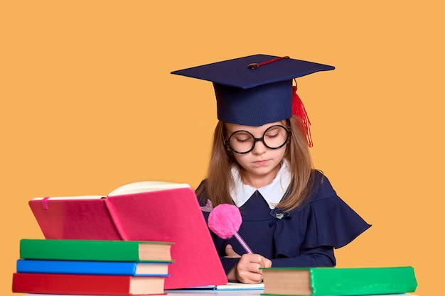 Curieuse écolière en tenue de graduation étudie avec des manuels scolaires