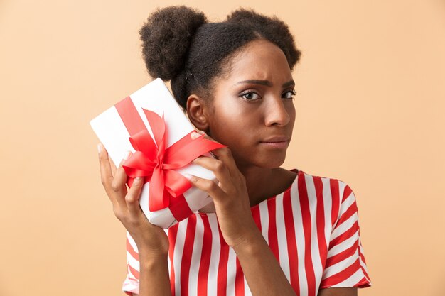 Curieuse femme afro-américaine dans des vêtements décontractés tenant une boîte-cadeau avec un arc rouge, isolé