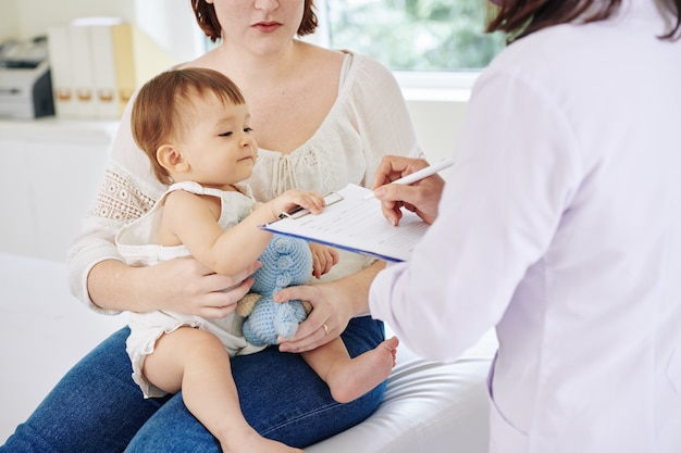 Curieuse petite fille regardant les antécédents médicaux que le pédiatre remplit en parlant à sa mère