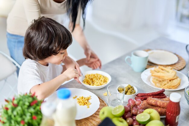 Curieux petit garçon hispanique aidant sa mère à préparer un repas pour le déjeuner, debout dans la cuisine à la maison ensemble. Maternité, enfants, concept de cuisine