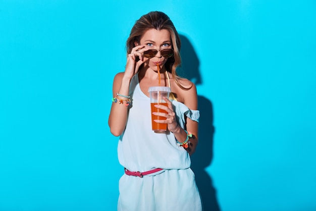 Curiosité. Jolie jeune femme regardant la caméra et ajustant ses lunettes en se tenant debout sur fond bleu