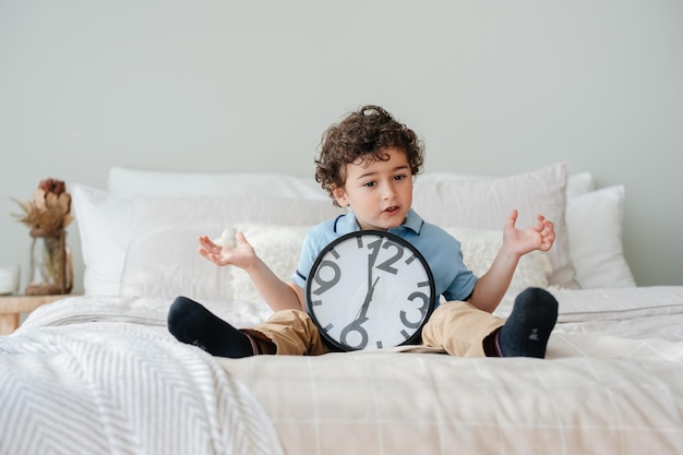 Curly beau garçon espagnol assis sur le lit avec une grande horloge murale écartant les mains dans l'expression d'incrédulité attendant que les parents passent du temps ensemble Concept de psychologie pour enfants Maquette temps de croissance des enfants