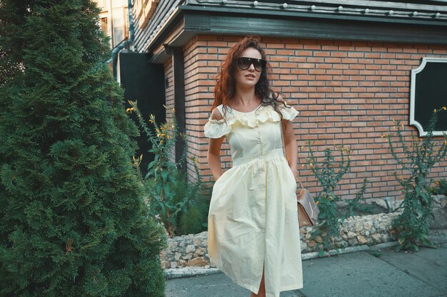 Curly belle femme dans une mince robe d'été marchant près du mur de briques