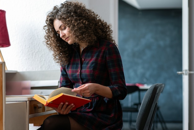 Curly brunette fille tenant un livre à la main et lisant, assis dans la chambre