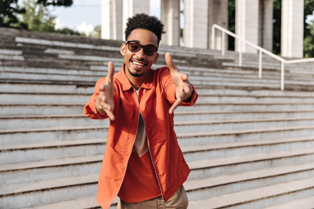 Curly jeune charmant homme à la peau foncée en tshirt veste orange et lunettes de soleil pointe vers la caméra et sourit près des escaliers à l'extérieur