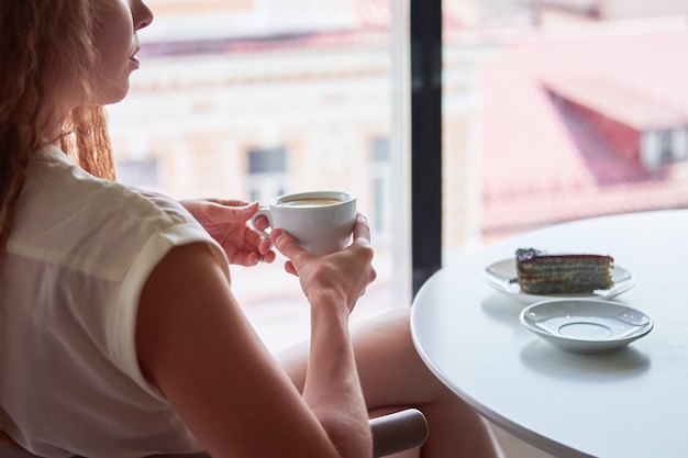Curly rousse maigre femme assise dans un café en face de la fenêtre avec une tasse de café et en observant le paysage urbain européen