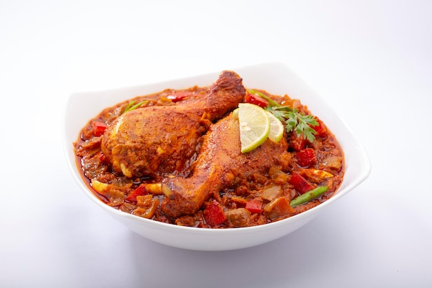 Curry de poulet indien ou masala de poulet, sauce épaisse au poulet épicé indien garnie de feuilles de coriandre, tranches de citron et disposées dans une vaisselle en céramique blanche ou un bol de service sur fond blanc, est