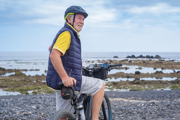 Cycliste barbu souriant homme âgé portant un casque debout le long de la plage de la mer regardant l'horizon au-dessus de l'eau