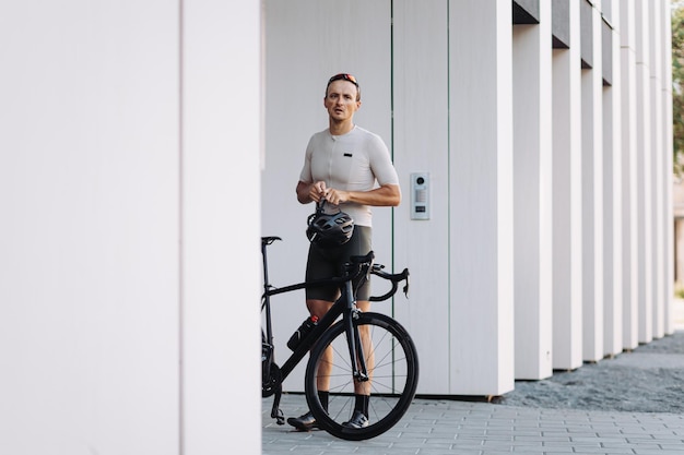 Cycliste caucasien épuisé debout près d'un vélo noir avec un casque de sécurité dans les mains Athlète masculin prenant une pause pendant l'exercice du matin en zone urbaine