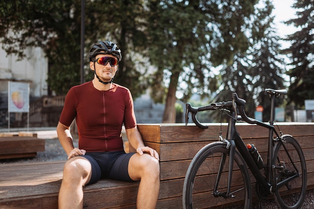 Cycliste caucasien en vêtements de sport, casque de sécurité et lunettes miroir se reposant après l'entraînement sur un banc en bois. Concept de personnes, de formation et de modes de vie.