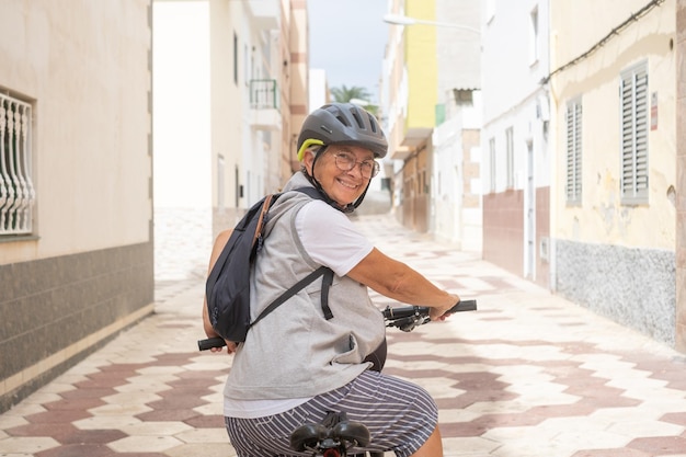 Cycliste joyeuse femme âgée dans la rue urbaine portant un casque et un sac à dos courant avec son vélo électrique Concept de mode de vie sain et de mobilité durable