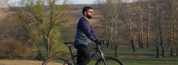 Cycliste en short et maillot sur un vélo semi-rigide moderne en carbone avec une fourche à suspension pneumatique debout sur une falaise sur fond de forêt printanière verte et fraîche