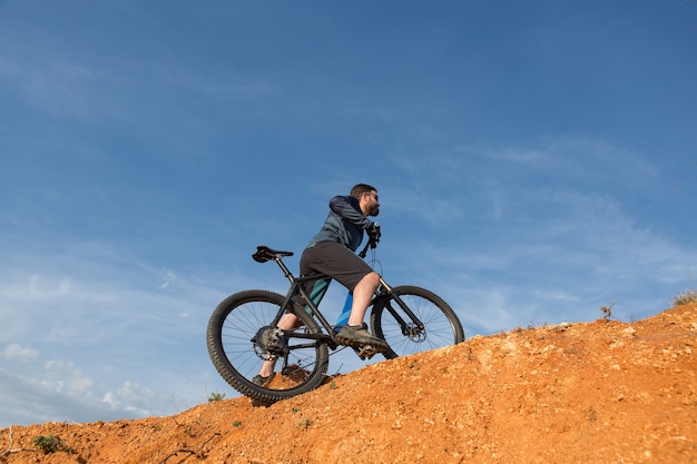 Cycliste en short et maillot sur un vélo semi-rigide moderne en carbone avec une fourche à suspension pneumatique debout sur une falaise sur fond de forêt printanière verte et fraîche