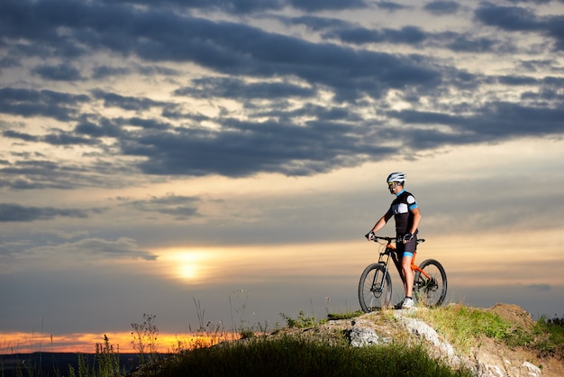 Cycliste avec un vélo repose sur une falaise dans les montagnes en profitant du soleil au coucher du soleil sous un ciel inhabituel avec de beaux nuages