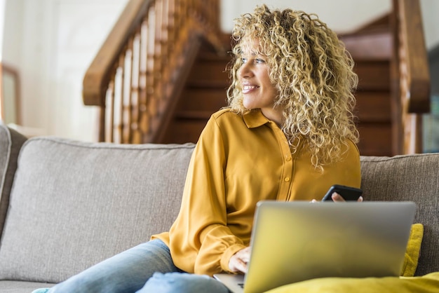 Dame adulte joyeuse avec de longs cheveux bouclés blonds travaille à la maison sur un ordinateur portable confortablement assis