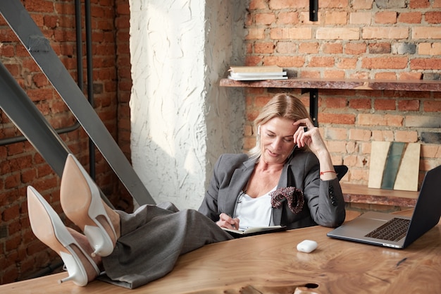 Dame d'affaires mature moderne contenu en gardant les pieds sur la table au bureau et en vérifiant les notes dans le planificateur