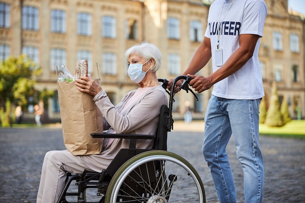 Dame âgée assise dans un fauteuil roulant avec un sac plein de nourriture. Bénévole non reconnu poussant les poignées